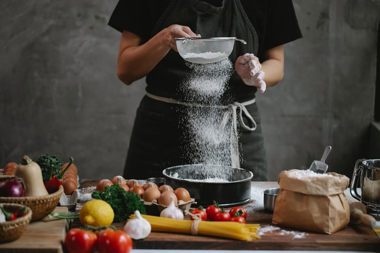 Crop unrecognizable cook adding flour into stainless baking dish while cooking meal with spaghetti cherry tomatoes garlic herbs and lemon